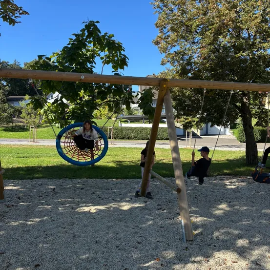 Drei Kinder spielen auf Schaukeln in einem Park. Eines schwingt auf einer Reifenschaukel, ein anderes auf einer traditionellen Schaukel und das dritte auf einer Reifenschaukel.