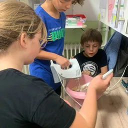 Drei Kinder sind in einer Küche, zwei benutzen einen Handmixer, um Zutaten in einer rosa Schüssel zu mischen.
