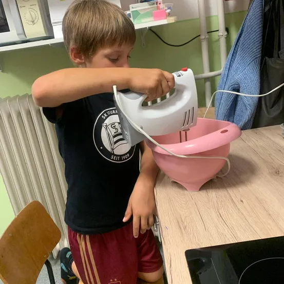 Ein kleiner Junge benutzt einen Handmixer, um Zutaten in einer rosa Schüssel auf einer Küchentheke zu mischen.