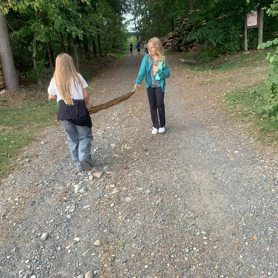Zwei Frauen gehen auf einem Kiesweg im Wald. Eine hält einen langen Stock, während die andere ihn am anderen Ende hält.