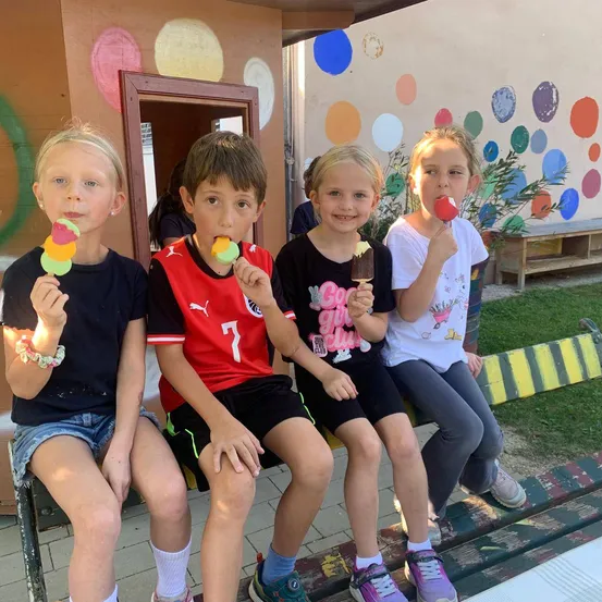 Fünf Kinder sitzen auf einer Bank und halten jeweils einen bunten Eis-Popsicle. Sie lächeln und posieren für ein Foto. Hinter ihnen ist eine Wand mit bunten Kreisen bemalt.