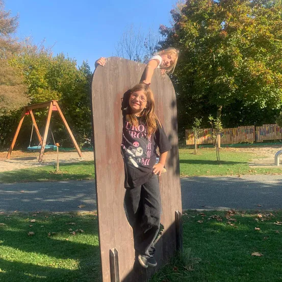 Zwei Mädchen stehen auf einem Holzbrett in einem Park. Ein Mädchen lehnt sich gegen das Brett, während das andere darauf steht. Hinter ihnen befindet sich eine Schaukel und ein farbenfroher Zaun.