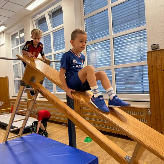 Zwei junge Jungen spielen auf einem Holzrutsche in einer Turnhalle. Ein Junge sitzt auf der Rutsche und der andere klettert hoch.