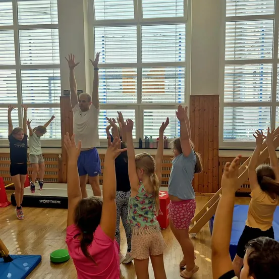 Eine Gruppe von Kindern in einem Fitnessstudio, die ihre Hände heben, während ein Mann in einem weißen Shirt auf einem Balancebalken steht.