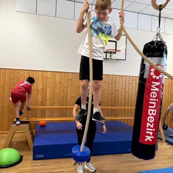 Ein junger Junge klettert in einem Fitnessstudio an einem Seil. Er trägt ein weißes T-Shirt mit einem farbenfrohen Design, schwarze Shorts und Turnschuhe.