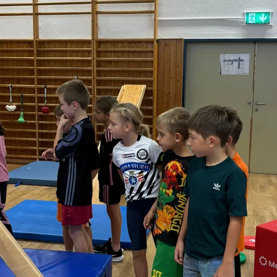 Eine Gruppe junger Kinder steht in einem Fitnessstudio, in Sportkleidung. Sie schauen auf etwas vor ihnen.