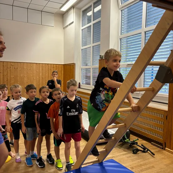 Eine Gruppe junger Jungen in einer Turnhalle, ein Junge klettert auf eine Holzleiter, während andere zusehen.