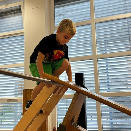 Ein kleiner Junge klettert auf eine Holzstruktur in einem Raum mit großen Fenstern.