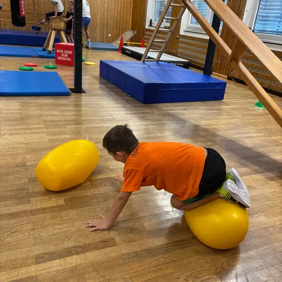 Ein junger Junge in einem orangefarbenen Shirt balanciert auf einem gelben Ball in einer Turnhalle.