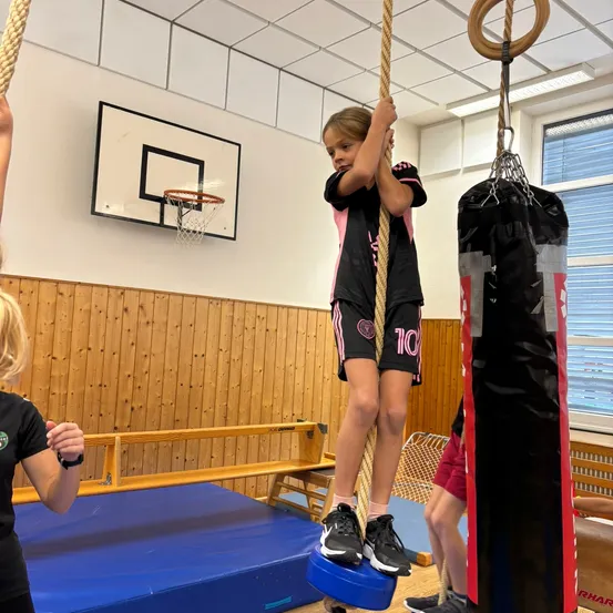 Ein junges Mädchen klettert in einem Fitnessstudio an einem Seil. Sie trägt ein schwarzes und rosafarbenes Sportoutfit mit der Nummer 10.