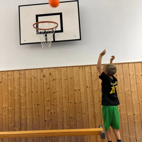 Ein junger Junge steht auf einer Holzplattform und wirft mit beiden Händen einen orangefarbenen Basketball in einen Basketballkorb, der an einer weißen Wand montiert ist.