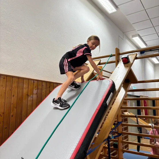 Ein junges Mädchen in einem schwarzen und rosa Sportoutfit klettert eine Holzrampe mit einem grünen Seil hinauf.
