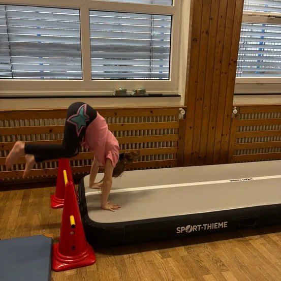 Ein junges Mädchen macht einen Handstand auf einer schwarzen und grauen aufblasbaren Matte auf einem Holzboden.