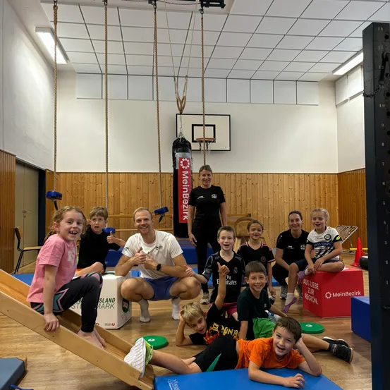 Eine Gruppe von Kindern und zwei Erwachsenen posiert für ein Foto in einem Fitnessstudio. Das Fitnessstudio hat Holzböden, weiße Decken und einen Basketballkorb.