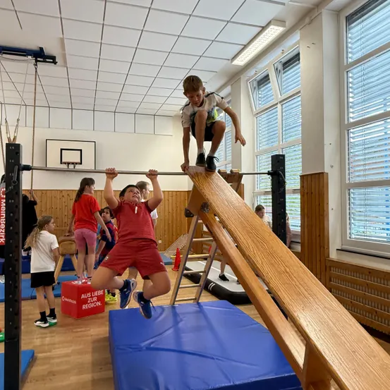 Zwei Kinder spielen auf einer Holzbalken in einer Turnhalle. Ein Kind springt vom Balken, während das andere hinaufklettert.