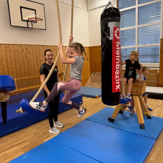 Ein junges Mädchen klettert in einem Fitnessstudio an einem Seil, wobei ein Boxsack an der Decke hängt.