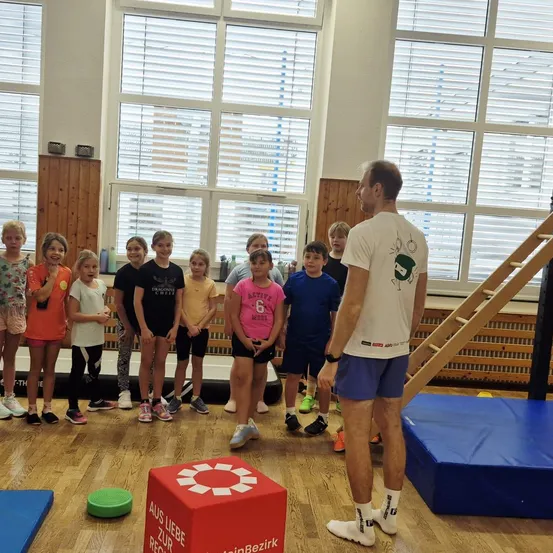 Eine Gruppe von Kindern und einem Mann sind in einem Fitnessstudio. Der Mann steht vor ihnen und sie alle schauen ihn an. Das Fitnessstudio hat Holzböden, einen Boxsack und eine blaue Matte.