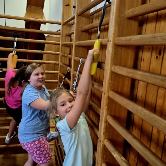 Drei junge Mädchen klettern in einem Fitnessstudio an einem Holzklettergerüst. Das Mädchen in der Mitte hält einen gelben Griff mit beiden Händen. Das Mädchen auf der linken Seite hält einen gelben Griff mit einer Hand. Das Mädchen auf der rechten Seite hält einen gelben Griff mit beiden Händen.