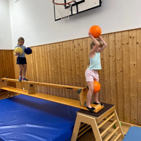 Zwei kleine Mädchen balancieren auf einem Holzbalken in einer Turnhalle. Eines hält einen gelben Ball und das andere einen orangefarbenen Ball.