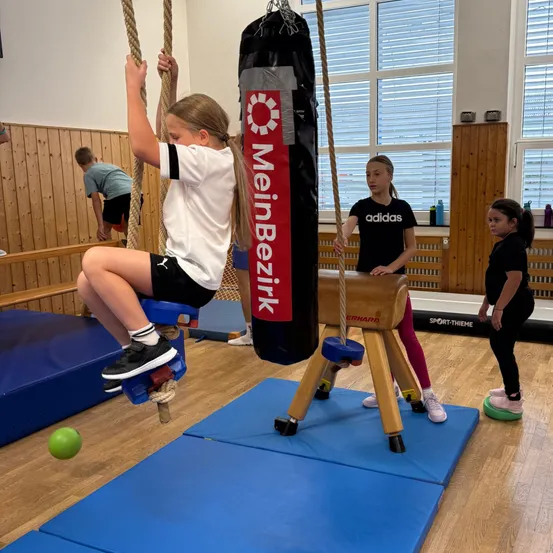Ein Mädchen klettert in einem Fitnessstudio an einem Seil, wobei ein Boxsack an der Decke hängt.