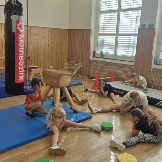 Eine Gruppe von Kindern macht Gymnastik auf dem Boden in einem Fitnessstudio. Sie sitzen auf blauen Matten und einige machen Übungen. Ein Boxsack hängt von der Decke.