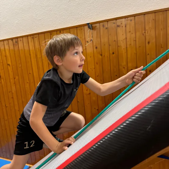 Ein junger Junge klettert mit einer grünen Seil in den Händen an einem großen aufblasbaren Rutschen hoch. Er trägt ein schwarz-graues Shirt mit der Nummer 12.