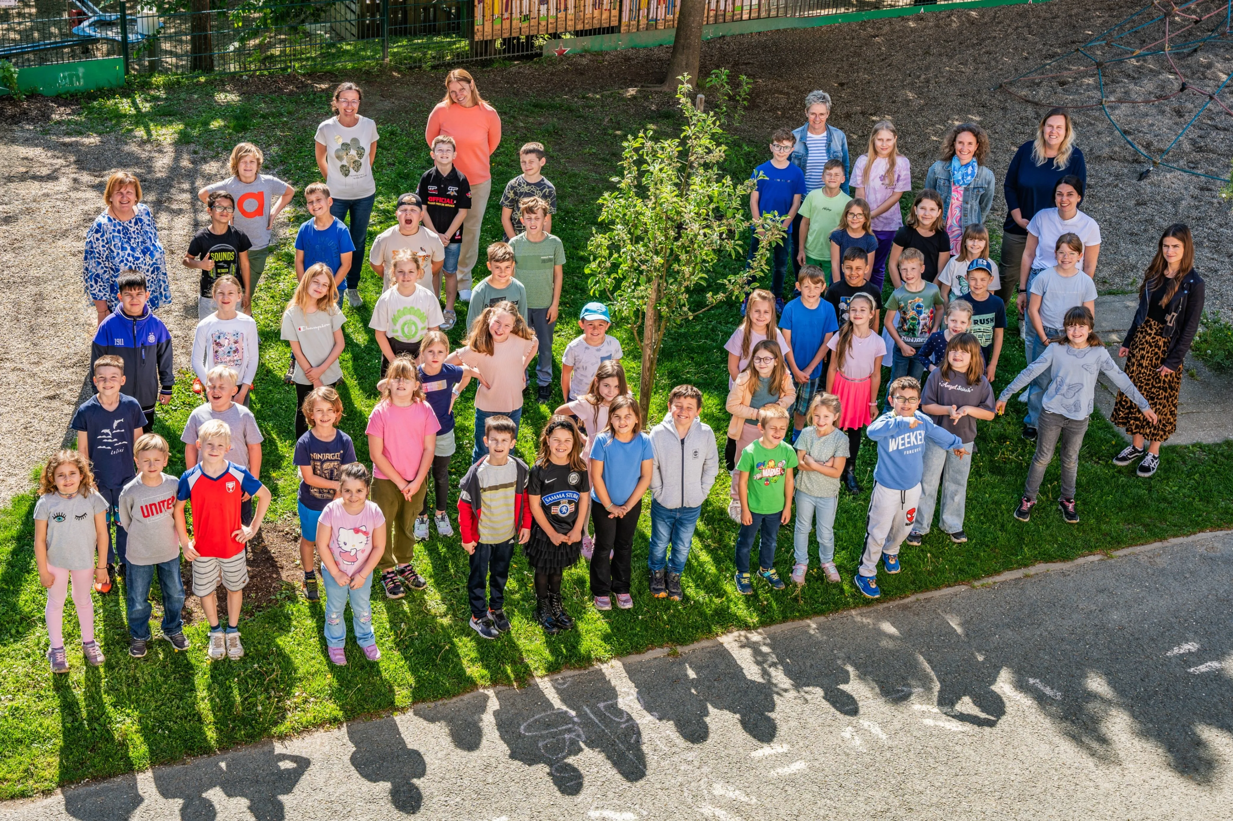 Eine Gruppe von Kindern und Erwachsenen versammelt sich auf einem Rasen, um für ein Foto zu lächeln. Der Boden hat weiße Kreide-Schrift. Im Hintergrund gibt es einen Zaun, einen Baum und einen Hügel.