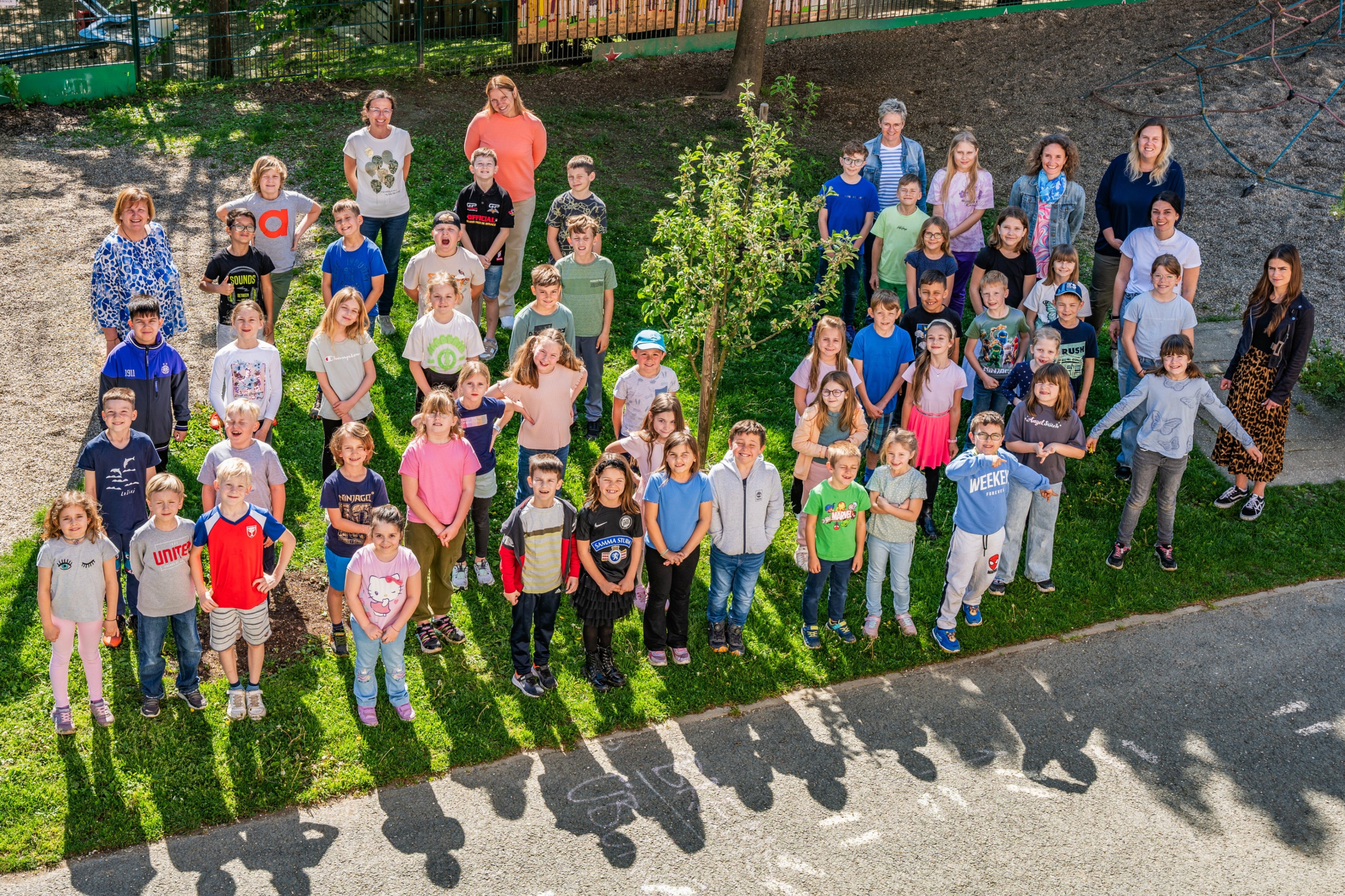 Eine Gruppe von Kindern und Erwachsenen versammelt sich auf einem Rasen, um für ein Foto zu lächeln. Der Boden hat weiße Kreide-Schrift. Im Hintergrund gibt es einen Zaun, einen Baum und einen Hügel.