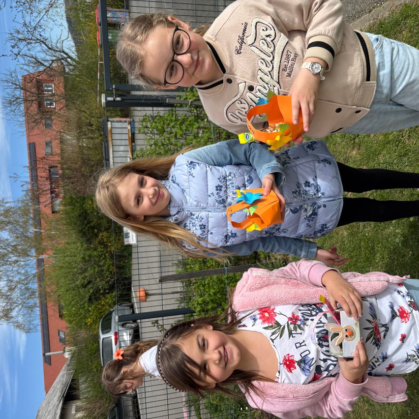 Drei junge Mädchen lächeln und posieren für ein Foto in einem eingezäunten Bereich. Das Mädchen in der Mitte hält einen kleinen orangefarbenen Korb, und die anderen beiden halten ein kleines Objekt. Hinter ihnen steht ein Gebäude mit einer Backsteinmauer und Glasfenstern.
