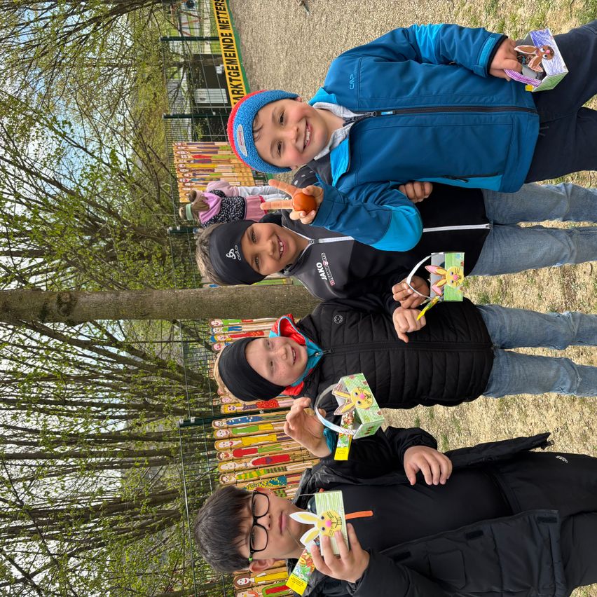 Vier Kinder in einem Park, zwei stehend, zwei sitzend, halten bunte Osterkörbe und lächeln. Bäume und ein gelber Zaun sind im Hintergrund zu sehen.