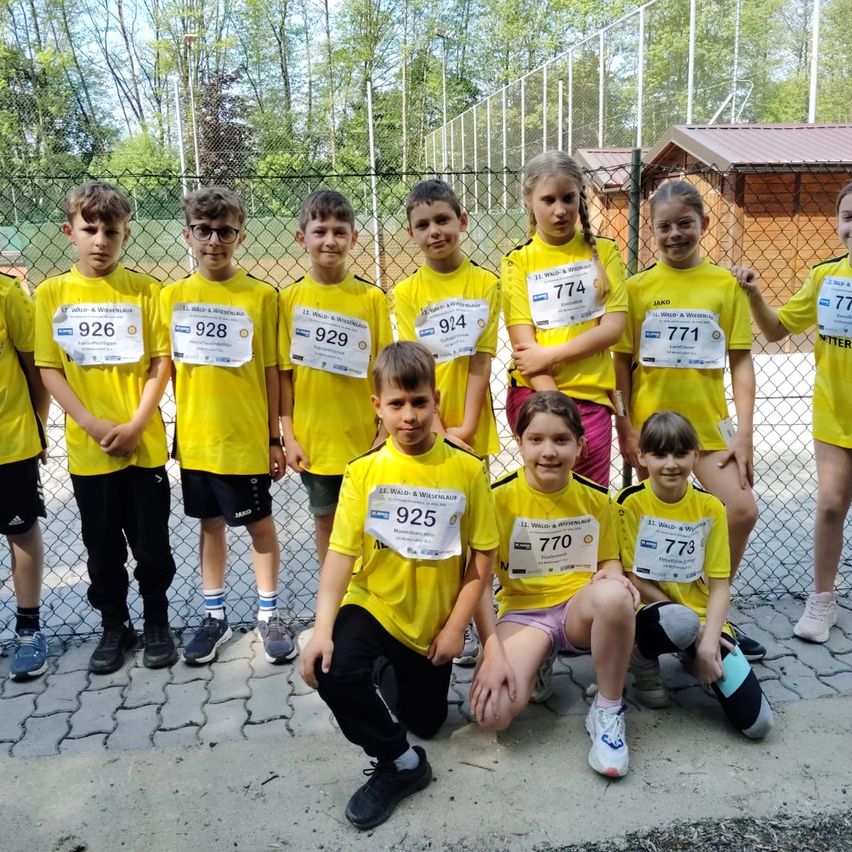 Eine Gruppe von Kindern, wahrscheinlich ein Sportteam, trägt gelbe Trikots und schwarze Shorts und posiert für ein Foto. Sie stehen und sitzen vor einem Zaun mit einem hölzernen Gebäude im Hintergrund.