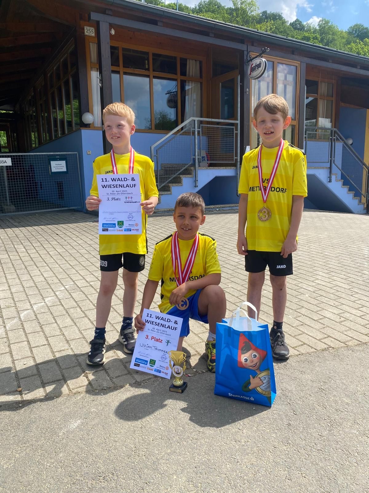 Drei junge Jungen in gelben Shirts und Shorts mit Medaillen um den Hals stehen auf einem Ziegelweg. Sie halten Urkunden und einen Pokal. Einer von ihnen hält eine blaue Tasche mit einem Comic-Charakter darauf. Hinter ihnen befindet sich ein Gebäude mit Treppen und einem Metallzaun.