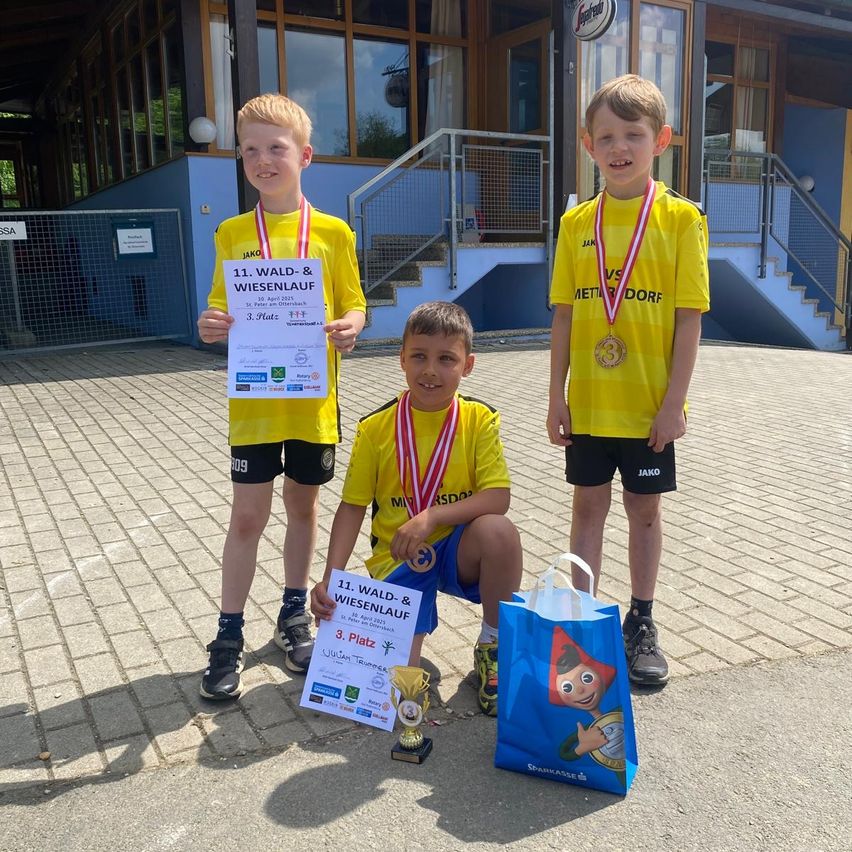 Drei junge Jungen in gelben Shirts und Shorts mit Medaillen um den Hals stehen auf einem Ziegelweg. Sie halten Urkunden und einen Pokal. Einer von ihnen hält eine blaue Tasche mit einem Comic-Charakter darauf. Hinter ihnen befindet sich ein Gebäude mit Treppen und einem Metallzaun.
