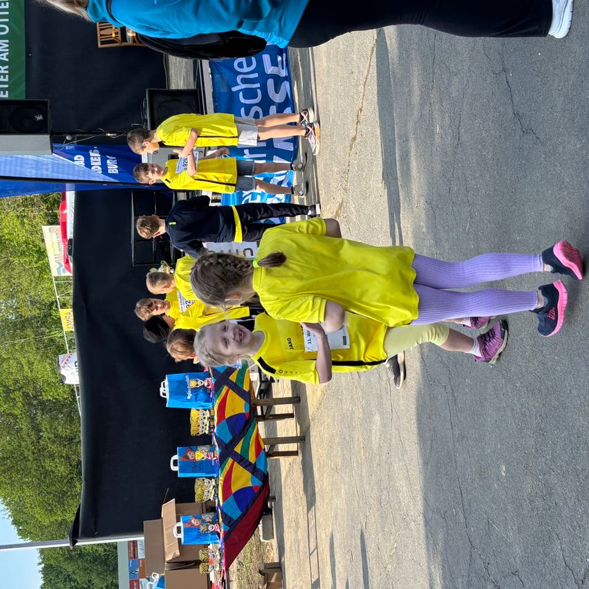 Kinder in gelben T-Shirts stehen in einer Reihe, wahrscheinlich an einer Veranstaltung teilnehmend. Sie tragen Turnschuhe und Leggings. Hinter ihnen stehen Personen in schwarzen T-Shirts, möglicherweise Organisatoren. Ein Tisch mit bunten Gegenständen ist in der Nähe, und ein Lautsprecher ist links aufgestellt.