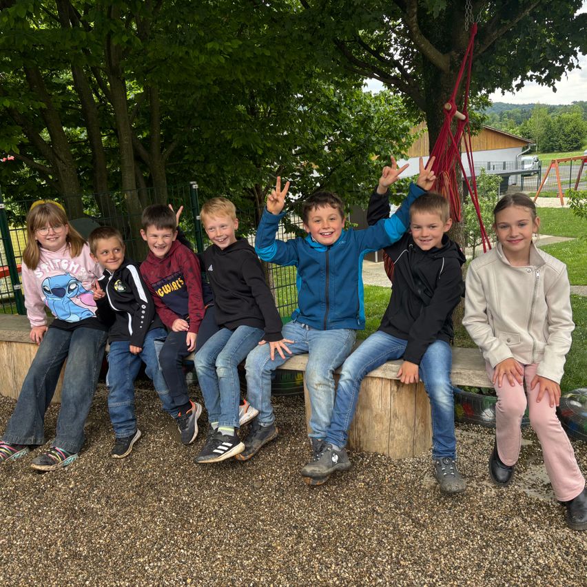 Eine Gruppe junger Kinder sitzt auf einer Holzbank in einem Park mit Bäumen und einer Schaukel im Hintergrund.