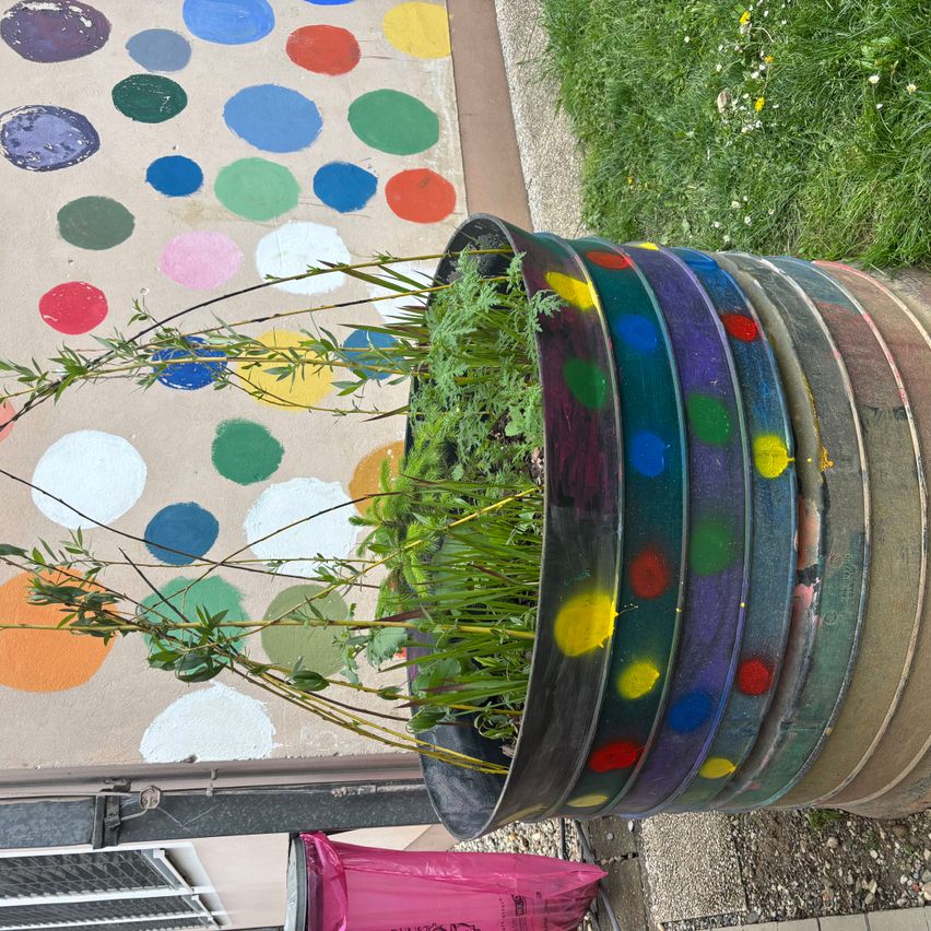 Ein bunter Blumentopf mit mehreren Ebenen, gefüllt mit Pflanzen, steht an einer Wand, die mit bunten Punkten verziert ist.