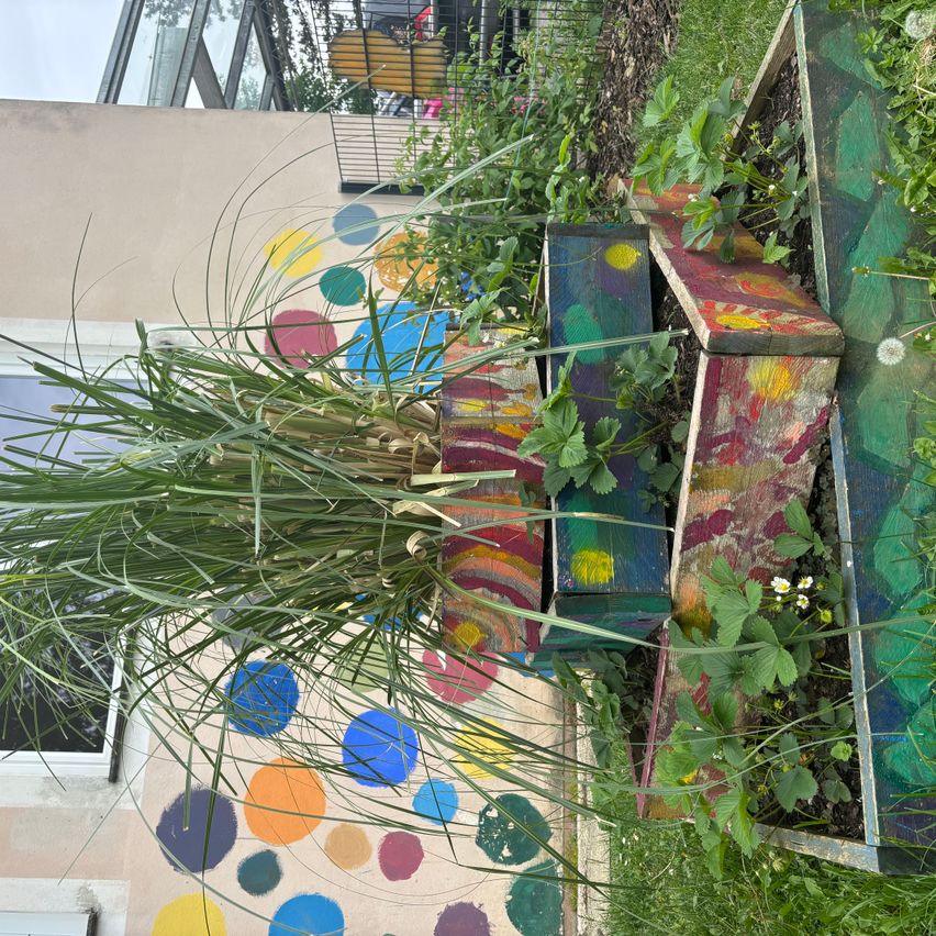 Ein Garten im Freien mit bunt bemalten Holzpflanzern. Ein hoher Strauch mit langen Blättern im Vordergrund. Die Wand dahinter ist mit bunten Punkten verziert.
