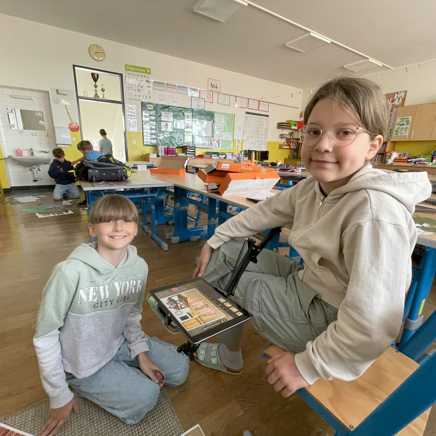 Zwei Kinder in einem Klassenzimmer mit einem Tisch dazwischen. Ein Kind trägt ein New York Sweatshirt und das andere eine Brille. Sie sitzen auf einem Stuhl und scheinen etwas auf einem Tablet zu tun.