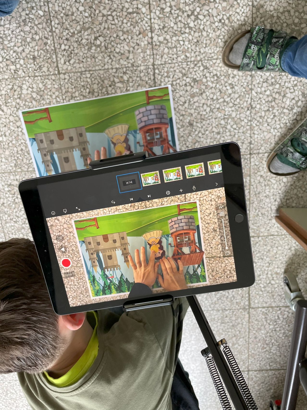 Ein Kind benutzt ein Tablet, um ein Bild zu zeichnen, mit einer Burgszene auf dem Bildschirm. Schuhe und ein Stativ sind im Hintergrund.