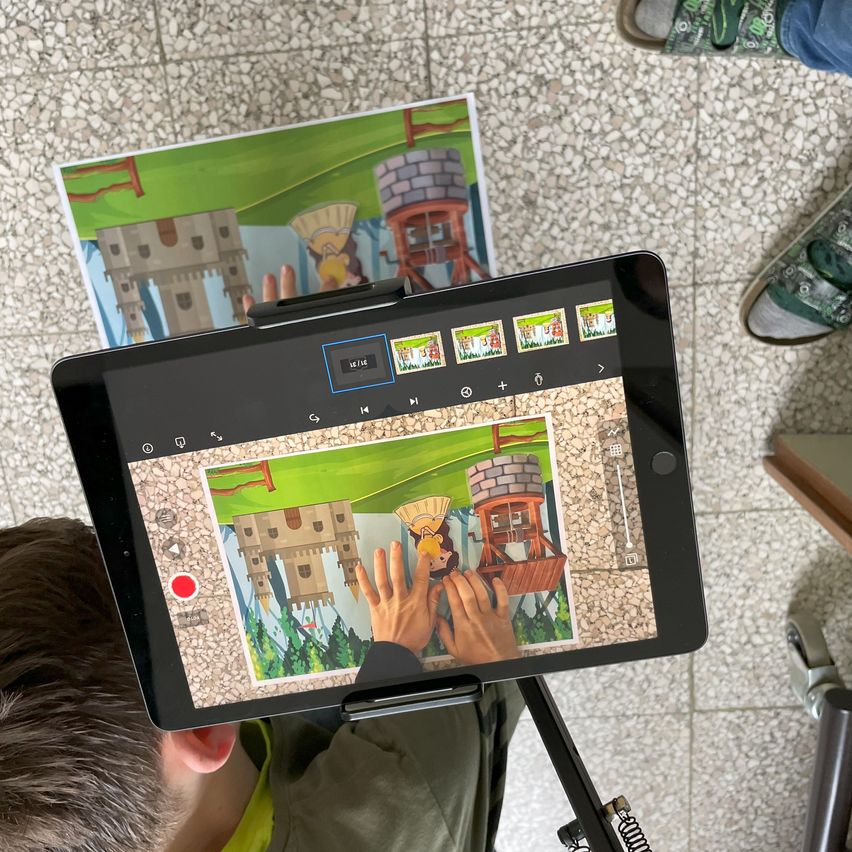 Ein Kind benutzt ein Tablet, um ein Bild zu zeichnen, mit einer Burgszene auf dem Bildschirm. Schuhe und ein Stativ sind im Hintergrund.