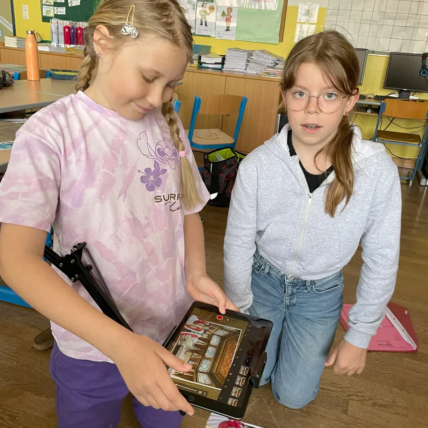 Zwei junge Mädchen sind in einem Klassenzimmer, eine kniet und die andere hält ein Tablet, beide konzentrieren sich auf den Bildschirm.