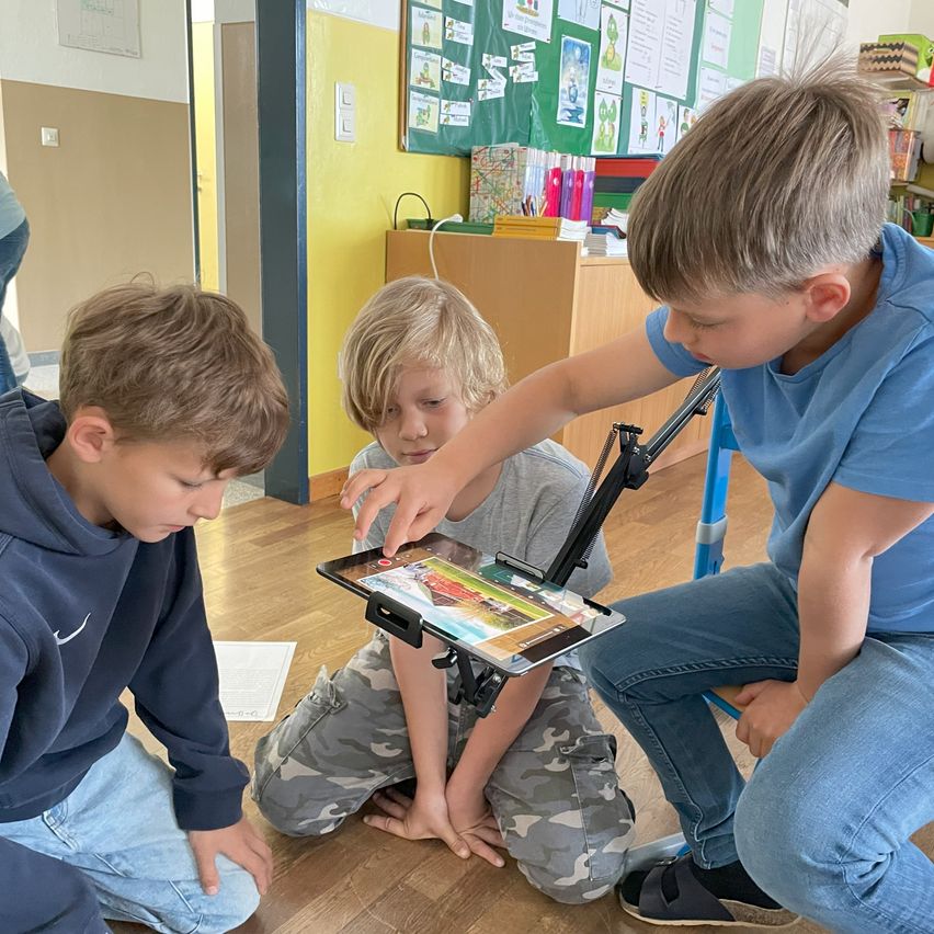 Drei Jungen sitzen auf dem Boden in einem Klassenzimmer und schauen auf ein Tablet. Ein Junge hält das Tablet, während die anderen aufmerksam zusehen. Hinter ihnen ist eine Pinnwand mit Papieren und Gegenständen gefüllt.