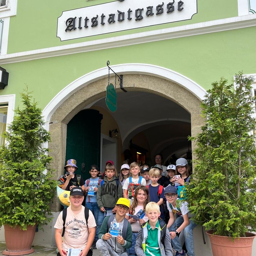 Eine Gruppe von Kindern, einige mit Hüten und Rucksäcken, posiert für ein Foto vor einem Gebäude mit der Aufschrift 'Altstadtgasse.' Der Eingangsbogen und die Topfpflanzen ergänzen die Szene.