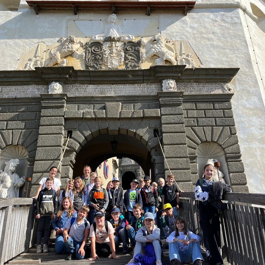 Eine Gruppe von Kindern und Erwachsenen steht auf einer Holzbrücke vor einem alten Steinbogen mit kunstvollen Schnitzereien und Statuen.