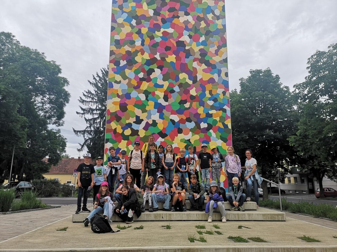 Eine Gruppe von Kindern und Erwachsenen posiert vor einer bunten, abstrakten Wandmalerei an einem sonnigen Tag. Sie stehen auf einer Betonplattform mit einem Geländer hinter ihnen.