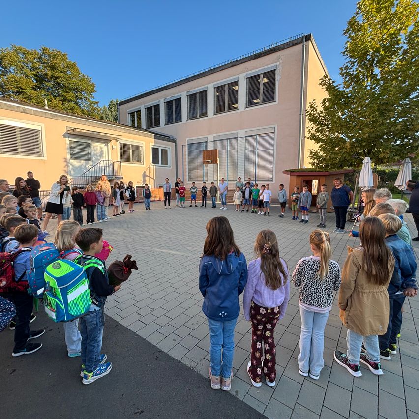 Eine Gruppe von Kindern und Erwachsenen steht in einem Kreis auf einem gepflasterten Bereich vor einem Gebäude. Das Gebäude hat mehrere Fenster und eine kleine Struktur mit einer Tür in der Nähe. Bäume und Regenschirme sind im Hintergrund sichtbar.