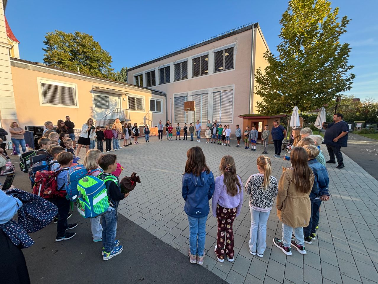 Eine Gruppe von Kindern und Erwachsenen steht in einem Kreis auf einem gepflasterten Bereich vor einem Gebäude. Das Gebäude hat mehrere Fenster und eine kleine Struktur mit einer Tür in der Nähe. Bäume und Regenschirme sind im Hintergrund sichtbar.