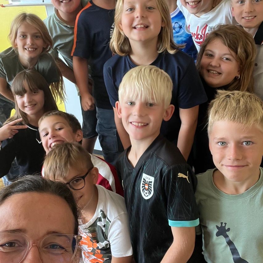 Eine Gruppe lächelnder Kinder, einige in Sporttrikots, posiert für ein Foto mit einer Frau in Brille. Sie sind in einem Raum mit gelber Wand.