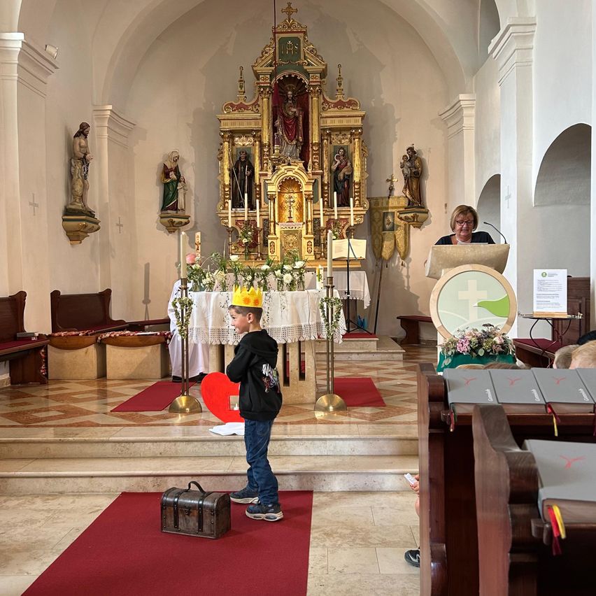 Ein junger Junge mit einer Krone steht in einer Kirche in der Nähe eines Altars, während eine Frau an einem Podium spricht. Der Altar ist mit Kerzen und Blumen geschmückt.