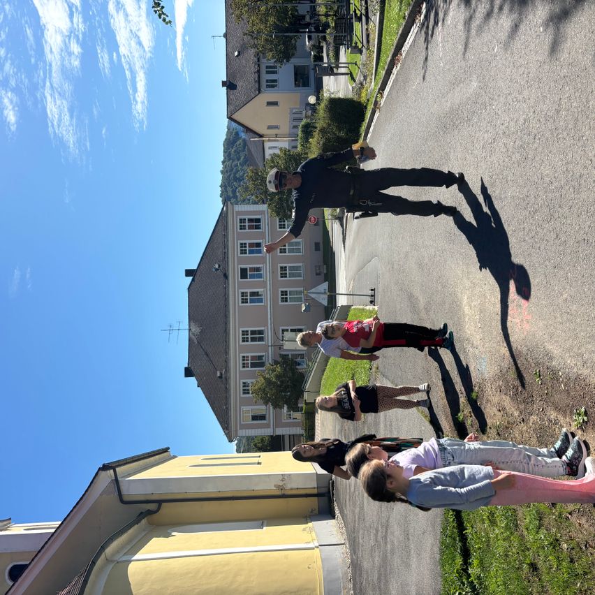 Eine Gruppe von Menschen, darunter ein Mann in Uniform, steht auf einer Straße mit Gebäuden und Bäumen im Hintergrund.